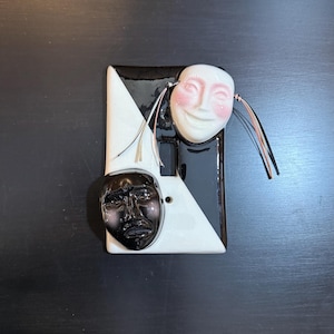May include: A decorative light switch cover featuring two ceramic masks. One mask is white with pink cheeks and a smiling expression, the other is black with a neutral expression. The cover has a black and white geometric design.