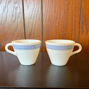 May include: Two white ceramic teacups with a blue geometric pattern around the rim. Each cup has a curved handle and a slightly flared shape. The cups are set against a wooden background.