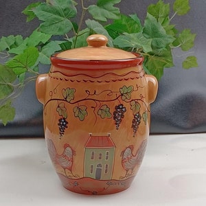 Peut inclure: Pot en céramique avec couvercle, décoré à la main. Le pot est de couleur beige avec un bord et des poignées rouges. Le motif comprend des vignes, des raisins, une maison verte et deux coqs. Le pot mesure environ 20 cm de haut.