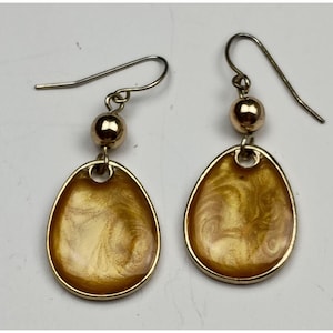 Peut inclure: Une paire de boucles d'oreilles dorées. Chaque boucle d'oreille présente un pendentif en forme de goutte d'eau avec un motif tourbillonnant et marbré dans des tons or et marron. Les boucles d'oreilles ont un accent de boule dorée et une fermeture à crochet.