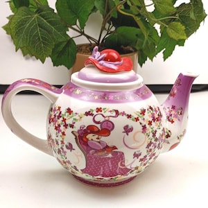 Puede incluir: Tetera de cerámica blanca con un diseño caprichoso que presenta a una mujer con un sombrero rojo, rodeada de motivos florales y mariposas. La tetera tiene un asa y un pico morados, y una tapa en forma de sombrero rojo. El diseño es detallado y colorido.