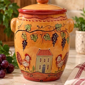 May include: A ceramic canister with a lid, featuring a hand-painted design of two roosters flanking a small house, set against a warm yellow background. The upper portion displays grapevines with purple clusters and green leaves.