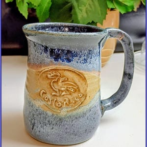 Puede incluir: Taza de cerámica hecha a mano con asa. La taza presenta un esmalte azul, beige y marrón con un acabado texturizado. Tiene un diseño en relieve con las palabras "Costa Rica" y un pájaro.