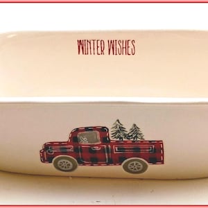 May include: White ceramic baking dish with red handles. The dish features a red plaid truck carrying Christmas trees, with the words "Winter Wishes" in red above the truck.