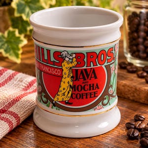 Op de afbeelding: Een witte keramische mok met het logo Hills Bros. San Francisco Java and Mocha Coffee. Het ontwerp bevat een figuur in een geel gewaad met patronen, rode en groene accenten en een gouden rand langs de boven- en onderkant van de mok.