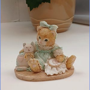 Peut inclure: Une figurine de chat roux portant une robe bleue et un nœud blanc, assise sur une base en bois avec une petite figurine de chat blanc à côté.
