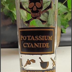 May include: A clear glass tumbler featuring a black and gold graphic of a skull and crossbones, the text POTASSIUM CYANIDE, small illustrations of a revolver, mortar and pestle, and a dagger, with the text NAME YOUR POISON at the base.