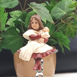 Könnte beinhalten: Eine Keramikfigur einer Frau, die ein Baby hält und ein weißes und weinrotes Outfit trägt. Die Figur sitzt auf einem durchsichtigen, blasenartigen Ständer, mit einem beigefarbenen Topf im Hintergrund und grünen Blättern.