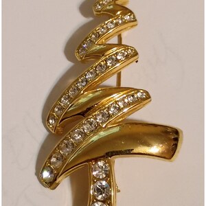 May include: A gold-tone metal brooch shaped like a stylized Christmas tree. The design features a swirling ribbon-like structure accented with clear, round, faceted rhinestones embedded along the edges and a vertical base set with three stones.