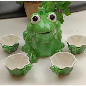Puede incluir: Un recipiente de cerámica verde con forma de rana con ojos grandes y una pequeña rana encima, acompañado de cuatro cuencos pequeños a juego. Los cuencos y el recipiente son verdes con interiores blancos, creando un conjunto caprichoso.