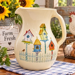 May include: A cream-colored ceramic pitcher featuring a painted design of four colorful birdhouses on poles behind a white picket fence. The birdhouses are yellow, blue, red, and white with various roof shapes, and a small brown bird sits on the fence.