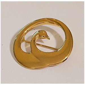 May include: A gold-toned brooch in the shape of a swan. The brooch features a stylized swan design with a curved body and a detailed head. The pin is a classic accessory.