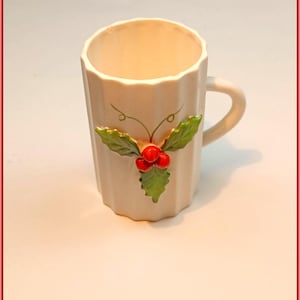 Puede incluir: Una taza de cerámica color crema con un diseño de acebo. La taza tiene un exterior acanalado y un asa curvada. El diseño de acebo presenta hojas verdes y bayas rojas, agregando un toque festivo. La taza es adecuada para bebidas calientes.