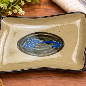 May include: A rectangular ceramic tray with a glossy beige speckled finish and a dark brown rim. The center features a dark green oval accent containing a stylized blue abstract graphic design. The tray has slightly curved, raised edges.