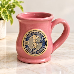 May include: A pink ceramic mug featuring a circular logo for The Original Pancake House. The logo includes a cartoon chef flipping a pancake, the text The Mosaic Directory Presents, The Original Pancake House, Since 1953, and Edina, Minnesota.