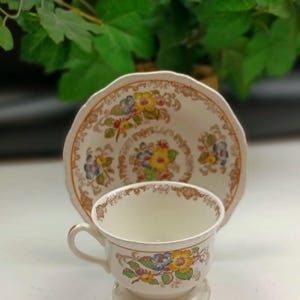 May include: A white teacup and saucer with a floral pattern in shades of yellow, orange, blue, and green. The floral pattern is surrounded by a brown border.