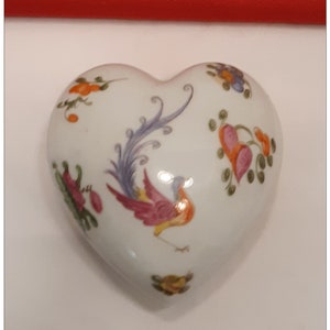 May include: A heart-shaped, white ceramic trinket box with hand-painted floral and bird designs in shades of red, pink, yellow, blue, and green. The box has a glossy finish and is likely used for storing small jewelry items.