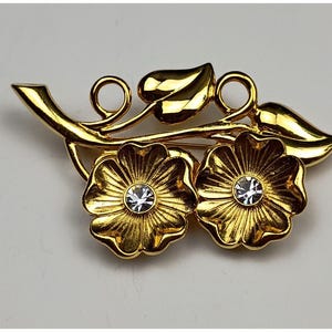 May include: A gold-toned floral brooch featuring two detailed flower blossoms with a sparkling crystal center. The brooch has a stem and leaf design, with a pin closure on the back.