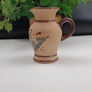 May include: A small, tan ceramic pitcher with a dark brown handle and rim. The pitcher features a floral design in blue, green, and brown. The pitcher is approximately 6 inches tall.