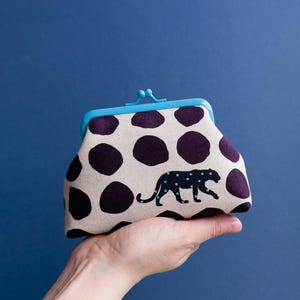 May include: A beige and purple polka-dot coin purse with a blue clasp. The purse features a black panther silhouette with blue spots. The purse is held in a hand against a blue background.
