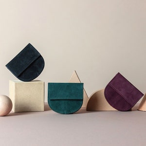 May include: Three half-circle shaped suede wallets in different colors: dark blue, teal green, and burgundy. The wallets are arranged on a light brown surface with geometric shapes behind them.