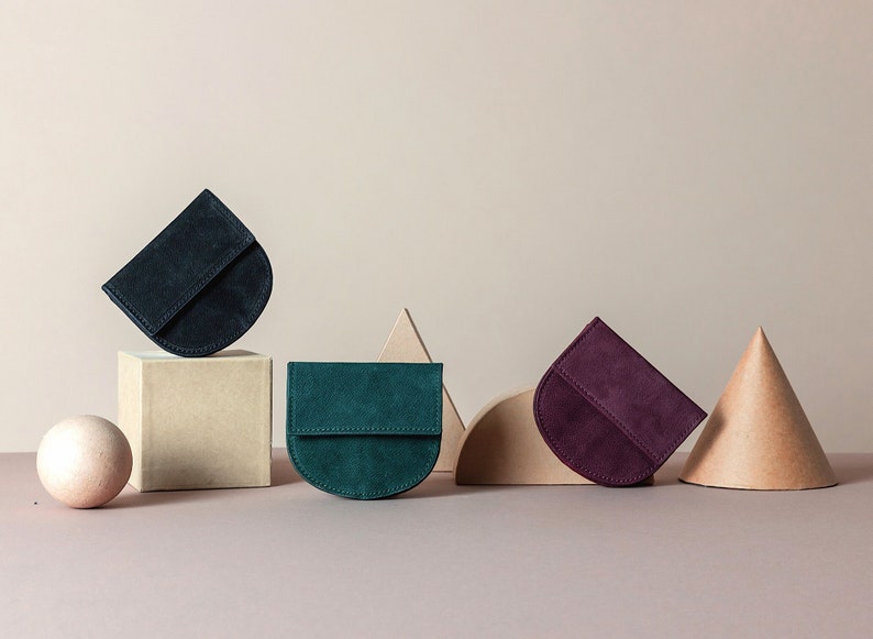 May include: Three half-circle shaped suede wallets in different colors: dark blue, teal green, and burgundy. The wallets are arranged on a light brown surface with geometric shapes behind them.