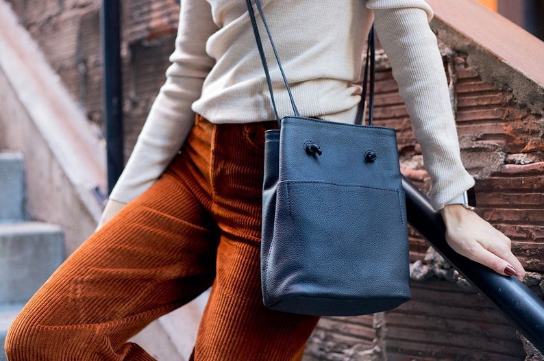 May include: A dark blue leather bucket bag with a drawstring closure and a long strap. The bag is worn over the shoulder of a person wearing a white sweater and orange corduroy pants.