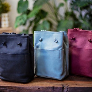 May include: Three small bucket bags in different colors: black, light blue, and burgundy. Each bag has a drawstring closure and a small front pocket.