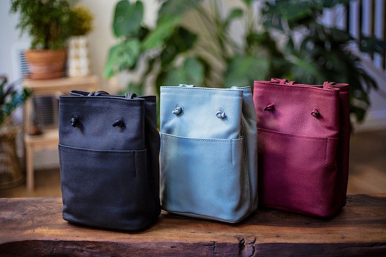 May include: Three small bucket bags in different colors: black, light blue, and burgundy. Each bag has a drawstring closure and a small front pocket.