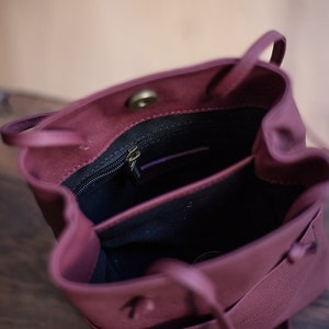 May include: A burgundy leather bucket bag with a drawstring closure and a zippered pocket inside. The bag is open and the interior is visible.
