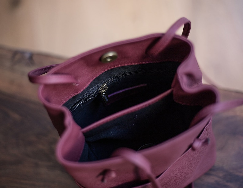 May include: A burgundy leather bucket bag with a drawstring closure and a zippered pocket inside. The bag is open and the interior is visible.