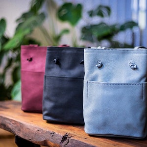 May include: Three leather bucket bags in different colors: burgundy, black, and light blue. The bags have a drawstring closure and a small pocket on the front.