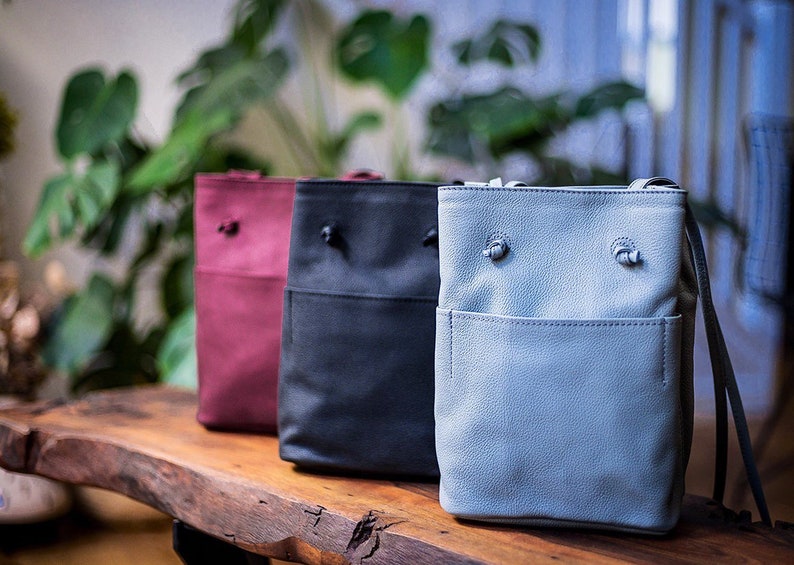 May include: Three leather bucket bags in different colors: burgundy, black, and light blue. The bags have a drawstring closure and a small pocket on the front.