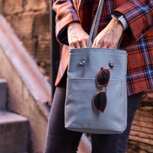 May include: A light gray leather tote bag with a top zipper closure and a front pocket. A pair of round sunglasses with brown frames are hanging from the bag.