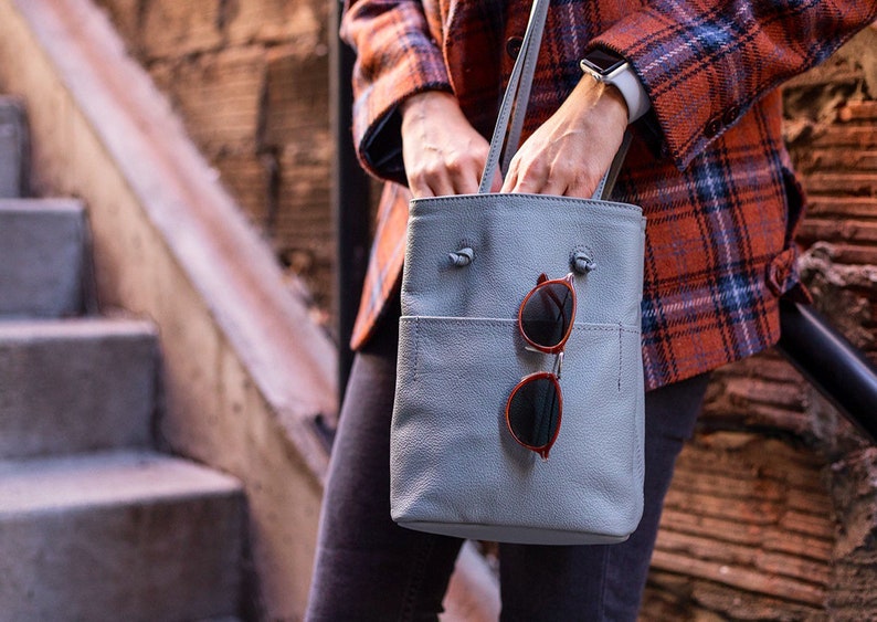 May include: A light gray leather tote bag with a top zipper closure and a front pocket. A pair of round sunglasses with brown frames are hanging from the bag.