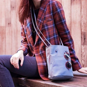 May include: A woman wearing a red and blue plaid blazer and dark blue jeans is sitting on a wooden bench. She is carrying a light blue tote bag with a pair of sunglasses attached to the front.