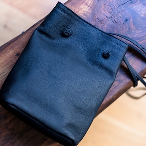 May include: A black leather drawstring bag with two black metal toggles. The bag is sitting on a wooden surface.