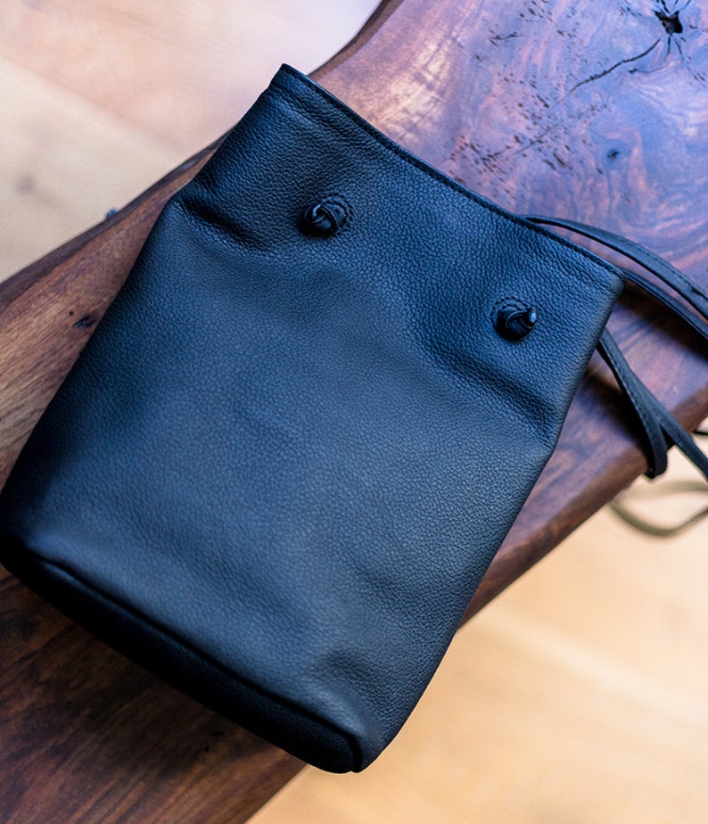 May include: A black leather drawstring bag with two black metal toggles. The bag is sitting on a wooden surface.