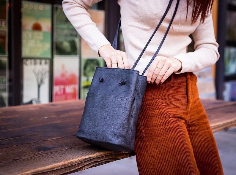 May include: A black leather bucket bag with a long strap. The bag is worn over the shoulder of a person wearing a white sweater and brown corduroy pants.
