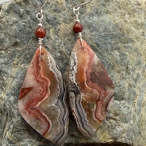 May include: A pair of dangle earrings featuring elongated, diamond-shaped slices of banded agate with swirling patterns of red, brown, gray, and white. Each earring is topped with a small, round, dark red bead and a silver-toned metal hook.