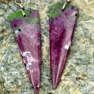 May include: A pair of dangle earrings featuring triangular-shaped, polished ruby and zoisite stones. The stones display a mottled pattern of deep magenta, vibrant green, and small black inclusions. Each stone is suspended from a silver-toned wire hook.