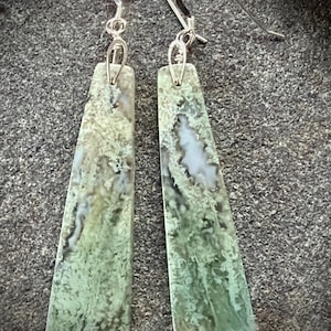 May include: A pair of dangle earrings featuring elongated, trapezoidal-shaped green moss agate stones with intricate white and dark green inclusions. The stones are suspended from silver-toned metal ear wires with a simple loop attachment mechanism.