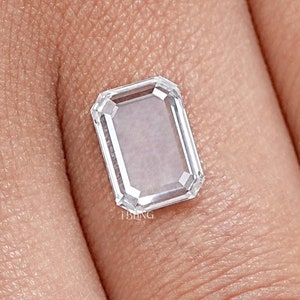Puede incluir: Una gema clara de corte rectangular con una superficie blanca y reflectante. La gema tiene una forma de esmeralda escalonada con una apariencia brillante y clara. La gema se coloca sobre un fondo de tono de piel.