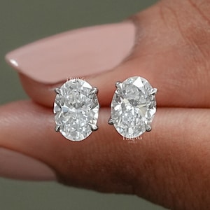 May include: A pair of oval-shaped diamond earrings set in a silver metal setting. The earrings are shown on a person's hand.