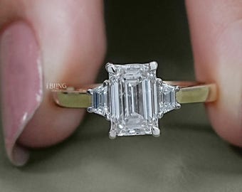 Puede incluir: Un anillo de oro con una piedra central de diamante talla esmeralda y dos piedras laterales más pequeñas. El anillo se sostiene entre dos dedos, mostrando el diseño y el brillo. El anillo está sobre un fondo neutro.