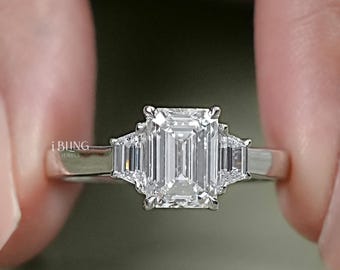 Puede incluir: Un anillo de plata con una piedra central de diamante de talla esmeralda y dos diamantes más pequeños de talla baguette a cada lado.