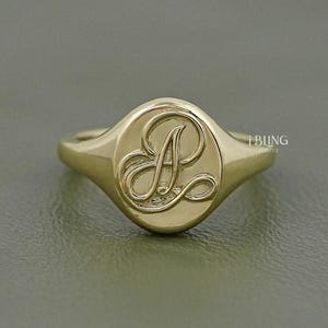 May include: A gold signet ring with a raised, cursive letter "A" inside an oval. The ring has a smooth, polished finish and a classic, elegant design. The ring is shown on a neutral background.