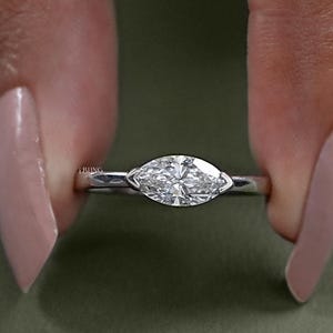 Puede incluir: Un anillo de diamantes con corte marquesa y una fina banda de plata. El diamante está sujeto entre dos garras. El anillo se sostiene entre dos dedos. Una joya clásica.