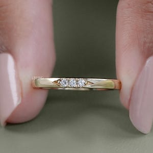 May include: A gold band ring with three small, clear gemstones set in a row. The ring is held between two fingers, showcasing its delicate design and polished finish. The ring's band has a subtle, geometric design.