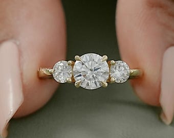 May include: A gold engagement ring featuring a large, round, clear central gemstone, with two smaller stones either side. The ring is held up by a person's fingers.